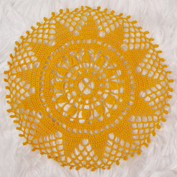 a mid-size, round crocheted doily made entirely with a bright golden yellow thread on a white faux fur background. the design looks like the sun; the center is a small circle with a cross through it, like an old window, with 13 small petals in a larger border around it. surrounding the sunflower-reminiscent motif in the center are a circle of 13 triangles pointing out toward the edges, like the sun. the spaces between the points are filled in with loose, lacy netting to complete the circle. 