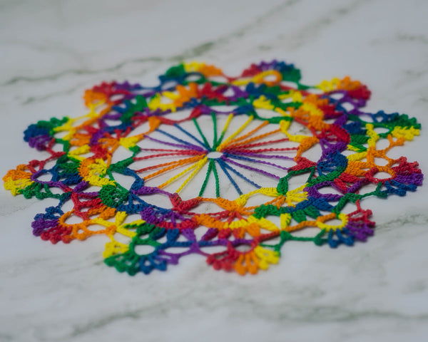 Doily done in three layers in variegated rainbow yarn. The center fans out like a ferris wheel into twelve points, the middle takes each point and attaches ‘containers’ to the spokes that themselves hold five smaller points, like people together. Each container there is topped with a circle from which the outer layer attaches, crowning each segment with nine-spoke fans. Set against a white marbled background.